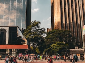 An urban street scene displays a bustling city environment with tall skyscrapers reflecting the sky and clouds. Trees provide greenery amidst the urban landscape, and a diverse group of people are walking and gathering on the street. There are various urban elements present, including street signs and building facades.