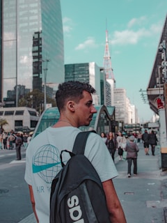 A young man with short dark hair carries a black backpack and wears a light blue t-shirt featuring a globe design with the words 'AIR FORCE.' He stands on a bustling city street with tall glass buildings, a red and white tower in the background, and numerous pedestrians walking around.