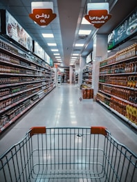 A supermarket aisle with products lined up on both sides. The view is from behind a shopping cart, looking down the aisle towards a distant checkout area. Overhead signs indicate sections for oil and pickles/jam. Shelves are neatly stocked with various bottled and packaged goods.