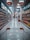 A supermarket aisle with products lined up on both sides. The view is from behind a shopping cart, looking down the aisle towards a distant checkout area. Overhead signs indicate sections for oil and pickles/jam. Shelves are neatly stocked with various bottled and packaged goods.