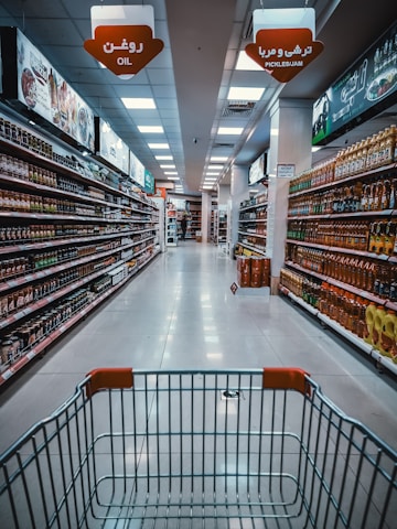 A supermarket aisle with products lined up on both sides. The view is from behind a shopping cart, looking down the aisle towards a distant checkout area. Overhead signs indicate sections for oil and pickles/jam. Shelves are neatly stocked with various bottled and packaged goods.