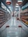 A supermarket aisle with products lined up on both sides. The view is from behind a shopping cart, looking down the aisle towards a distant checkout area. Overhead signs indicate sections for oil and pickles/jam. Shelves are neatly stocked with various bottled and packaged goods.