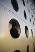 A black pair of over-ear headphones is hanging on a modern structure with a series of large circular cutouts. The surface is metallic and reflective, with visible fasteners that add an industrial feel.