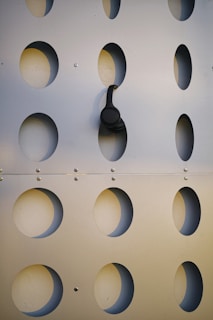 A perforated metal panel with evenly spaced oval cutouts. A pair of black headphones is hanging from one of the oval openings, creating a contrast against the metallic surface.