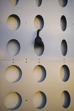 A perforated metal panel with evenly spaced oval cutouts. A pair of black headphones is hanging from one of the oval openings, creating a contrast against the metallic surface.