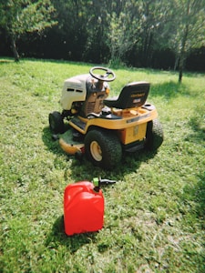 A yellow ride-on lawn mower is parked on a grassy lawn, surrounded by lush greenery. In front of the mower, there is a red fuel canister resting on the grass. The setting appears to be a sunny day with light filtering through the trees in the background.