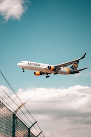 An airplane with the wording 'Condor' is flying in a clear blue sky, above a chain-link fence topped with barbed wire. The aircraft features yellow and navy blue accents.