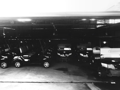 Multiple golf carts are parked in a dimly lit garage. The vehicles are aligned in a neat row, and the lighting creates reflective highlights on their surfaces.