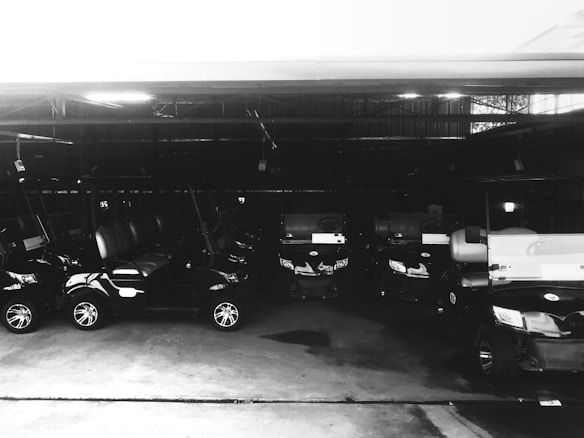 Multiple golf carts are parked in a dimly lit garage. The vehicles are aligned in a neat row, and the lighting creates reflective highlights on their surfaces.
