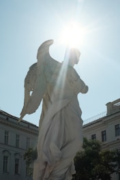A white stone statue of an angel with prominent wings stands in an urban setting. The sun shines brightly from behind the statue's head, creating a halo effect with sun rays radiating outward. The buildings in the background have classic architectural features, and there are leafy green trees surrounding the scene.