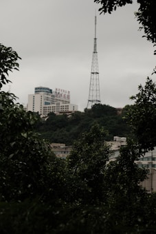 A tall communication tower stands against a cloudy sky, surrounded by lush green foliage. In the background, modern buildings with signage can be seen partially obscured by trees.