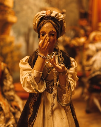 A detailed marionette puppet dressed in elaborate clothing, including a headscarf, necklace, and bracelets. The puppet's hand is raised near its face, and it is positioned against a blurred background that suggests a setting rich in warm, golden tones and soft lighting.