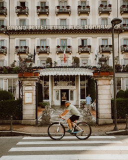 A grand hotel facade features multiple windows with small balconies adorned with potted plants. The entrance is ornate, flanked by statues and lush greenery. A cyclist passes by on a zebra crossing in front of the hotel, adding movement to the scene.