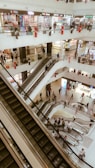 A multi-level shopping mall interior with several escalators and people moving about. Brightly lit stores with a variety of colorful signs line the upper floors. The overall architecture is modern with white walls and open spaces. Shoppers of varying ages and styles are seen ascending and descending the escalators.