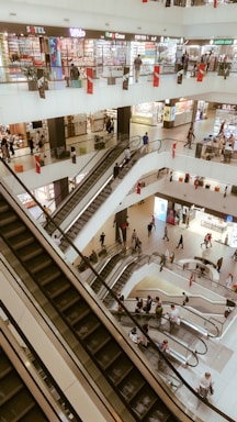 A multi-level shopping mall interior with several escalators and people moving about. Brightly lit stores with a variety of colorful signs line the upper floors. The overall architecture is modern with white walls and open spaces. Shoppers of varying ages and styles are seen ascending and descending the escalators.