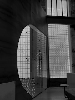 A modern architectural interior featuring a glass wall made of square panels that allows light to filter through. The wall is adjacent to a large dark wall with a curved cutout. The space includes a glass door with metal handles and a small round table with a flat top. The interior has a minimalistic and industrial design with clean lines.