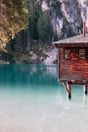 A rustic wooden cabin sits on stilts over a calm, turquoise lake. Surrounding the lake are lush evergreen trees and steep rock cliffs, creating a serene and picturesque setting.