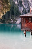 A rustic wooden cabin sits on stilts over a calm, turquoise lake. Surrounding the lake are lush evergreen trees and steep rock cliffs, creating a serene and picturesque setting.