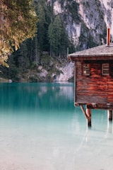 A rustic wooden cabin sits on stilts over a calm, turquoise lake. Surrounding the lake are lush evergreen trees and steep rock cliffs, creating a serene and picturesque setting.