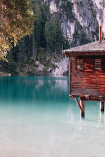 A rustic wooden cabin sits on stilts over a calm, turquoise lake. Surrounding the lake are lush evergreen trees and steep rock cliffs, creating a serene and picturesque setting.