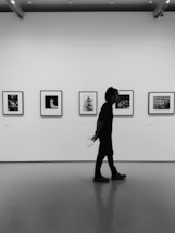 A person walking past a gallery wall lined with framed black-and-white photographs, holding a piece of paper behind their back. The gallery has a modern, minimalist design with a smooth floor and spotlights on the ceiling.