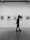 A person walking past a gallery wall lined with framed black-and-white photographs, holding a piece of paper behind their back. The gallery has a modern, minimalist design with a smooth floor and spotlights on the ceiling.