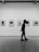 A person walking past a gallery wall lined with framed black-and-white photographs, holding a piece of paper behind their back. The gallery has a modern, minimalist design with a smooth floor and spotlights on the ceiling.