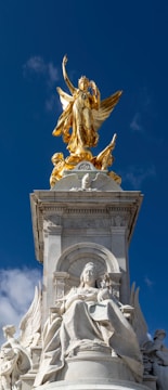 A prominent statue with a golden figure standing triumphantly on top of a stone pedestal. Beneath it, detailed carvings of historical figures are intricately crafted in alabaster-colored stone. The background shows a clear blue sky with sparse clouds.