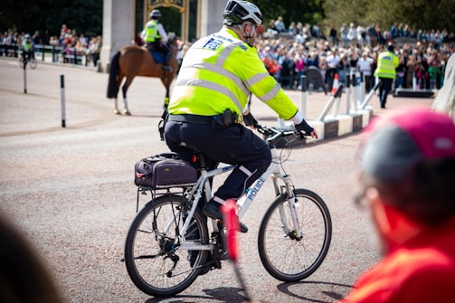 A police officer wearing a high-visibility jacket and helmet is riding a bicycle. The scene takes place in a crowded area with spectators observing from the background, while other officers, including one on horseback, are also visible.