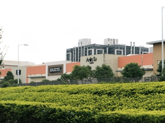 A shopping center with several retail store signs, including 'HAZZYS' and 'Anita Su'. There are modern buildings in the background and a neatly trimmed hedge and trees in the foreground. The sky is overcast, and there are streetlights along the path.