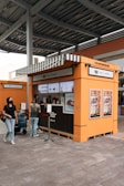 A kiosk with an orange and brown exterior is located beneath an industrial-style roof. The kiosk features a striped awning and is equipped with digital screens displaying menu items. Several posters are displayed on the kiosk's sides. A small queue of people wait to be served, with one person already interacting with the staff inside. A person is seen walking by, wearing casual attire.
