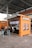 A kiosk with an orange and brown exterior is located beneath an industrial-style roof. The kiosk features a striped awning and is equipped with digital screens displaying menu items. Several posters are displayed on the kiosk's sides. A small queue of people wait to be served, with one person already interacting with the staff inside. A person is seen walking by, wearing casual attire.