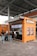 A kiosk with an orange and brown exterior is located beneath an industrial-style roof. The kiosk features a striped awning and is equipped with digital screens displaying menu items. Several posters are displayed on the kiosk's sides. A small queue of people wait to be served, with one person already interacting with the staff inside. A person is seen walking by, wearing casual attire.