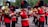A marching band dressed in red uniforms with black and gold accents, performing in an outdoor setting. The musicians are playing brass instruments like trombones and sousaphones, and they wear tall black hats with red plumes. Small flags are attached to their instruments.