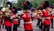 A marching band dressed in red uniforms with black and gold accents, performing in an outdoor setting. The musicians are playing brass instruments like trombones and sousaphones, and they wear tall black hats with red plumes. Small flags are attached to their instruments.