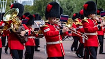 A marching band dressed in red uniforms with black and gold accents, performing in an outdoor setting. The musicians are playing brass instruments like trombones and sousaphones, and they wear tall black hats with red plumes. Small flags are attached to their instruments.