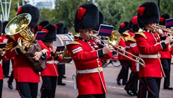 A marching band dressed in red uniforms with black and gold accents, performing in an outdoor setting. The musicians are playing brass instruments like trombones and sousaphones, and they wear tall black hats with red plumes. Small flags are attached to their instruments.