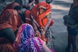 A group of women dressed in vibrant traditional clothing gather by a body of water. One woman, wearing an orange sari, is focused on performing a ritual with a small idol and a pot, possibly involving prayer or offerings. The women are adorned with bangles, and their attire features intricate patterns. The scene is intimate, depicting a sense of community and cultural tradition.