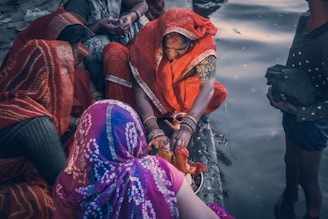 A group of women dressed in vibrant traditional clothing gather by a body of water. One woman, wearing an orange sari, is focused on performing a ritual with a small idol and a pot, possibly involving prayer or offerings. The women are adorned with bangles, and their attire features intricate patterns. The scene is intimate, depicting a sense of community and cultural tradition.