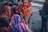 A group of women dressed in vibrant traditional clothing gather by a body of water. One woman, wearing an orange sari, is focused on performing a ritual with a small idol and a pot, possibly involving prayer or offerings. The women are adorned with bangles, and their attire features intricate patterns. The scene is intimate, depicting a sense of community and cultural tradition.
