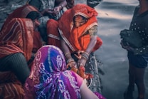 A group of women dressed in vibrant traditional clothing gather by a body of water. One woman, wearing an orange sari, is focused on performing a ritual with a small idol and a pot, possibly involving prayer or offerings. The women are adorned with bangles, and their attire features intricate patterns. The scene is intimate, depicting a sense of community and cultural tradition.