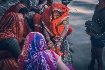 A group of women dressed in vibrant traditional clothing gather by a body of water. One woman, wearing an orange sari, is focused on performing a ritual with a small idol and a pot, possibly involving prayer or offerings. The women are adorned with bangles, and their attire features intricate patterns. The scene is intimate, depicting a sense of community and cultural tradition.