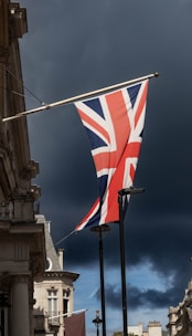 A Union Jack flag is prominently displayed on a pole against a dramatic dark sky with a building featuring classical architecture in the foreground.