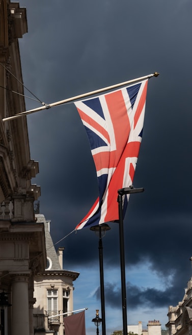 A Union Jack flag is prominently displayed on a pole against a dramatic dark sky with a building featuring classical architecture in the foreground.
