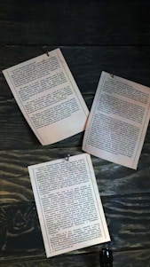 Three sheets of printed text are pinned to a wooden background. The pages are arranged in a slightly scattered manner, each held in place by small metallic tacks. The wood grain is visibly detailed, with a dark stain, creating a vintage or rustic feel.