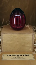 A decorative, dark red egg with intricate designs rests on a wooden stand surrounded by straw. Below the stand, a small label with text in a Cyrillic script provides information about the egg.