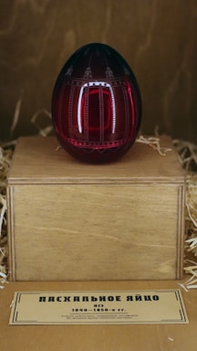 A decorative, dark red egg with intricate designs rests on a wooden stand surrounded by straw. Below the stand, a small label with text in a Cyrillic script provides information about the egg.