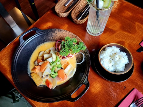A delicious-looking meal is served in a black bowl with a creamy orange-colored curry filled with vegetables such as carrots, cabbage, and broccoli, garnished with fresh parsley. Next to it, a bowl of white rice sprinkled with sesame seeds is placed. A glass of iced drink with a slice of lime and a straw accompanies the meal, all set on a wooden table with cutlery nearby.