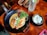 A delicious-looking meal is served in a black bowl with a creamy orange-colored curry filled with vegetables such as carrots, cabbage, and broccoli, garnished with fresh parsley. Next to it, a bowl of white rice sprinkled with sesame seeds is placed. A glass of iced drink with a slice of lime and a straw accompanies the meal, all set on a wooden table with cutlery nearby.
