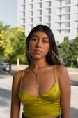 A young woman with long dark hair standing outdoors, wearing a bright lime green camisole top and a delicate necklace. The background features a modern building with a facade consisting of repetitive geometric patterns and lush green trees on a sunny day.